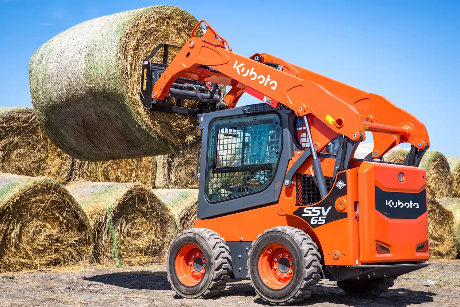 Kubota SSV65 skid steer lifting a round hay bale beside a stack of bales